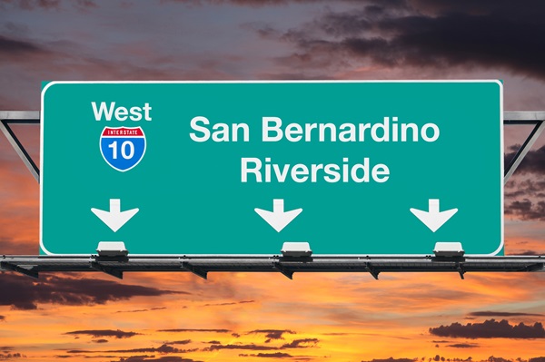 Green highway sign on Interstate 10 directing drivers west toward San Bernardino and Riverside with a sunset sky in the background.