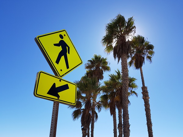 Yellow pedestrian crossing sign with an arrow pointing left, set against a clear blue sky and tall palm trees in Los Angeles.